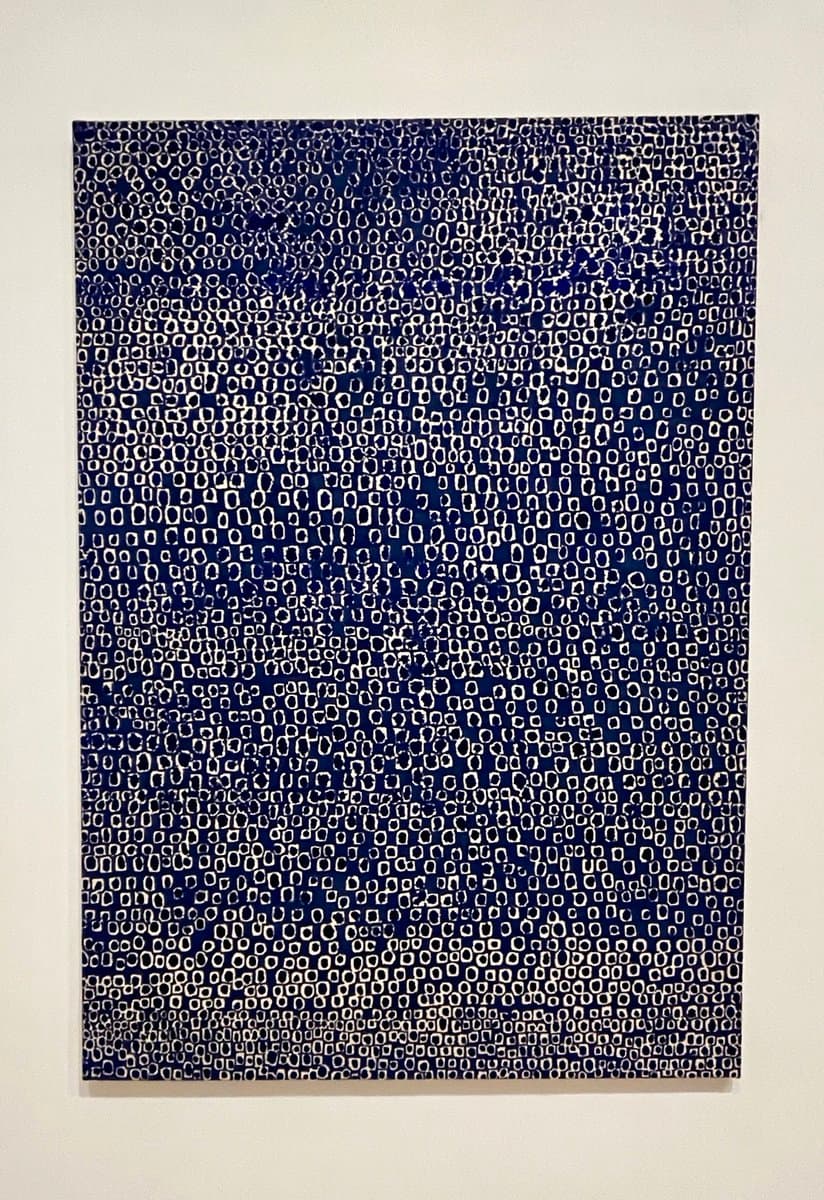 A vertical abstract painting featuring a dense, repetitive pattern of small white and black shapes on a dark blue background, hanging on a plain white wall.