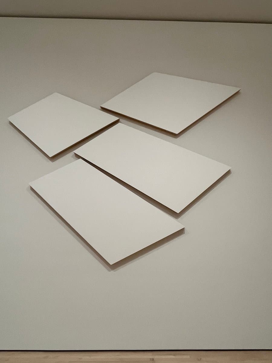 A minimalist art installation featuring four plain white rectangular panels mounted on a light gray wall. The panels are arranged in a staggered, diagonal formation, creating a simple geometric composition.