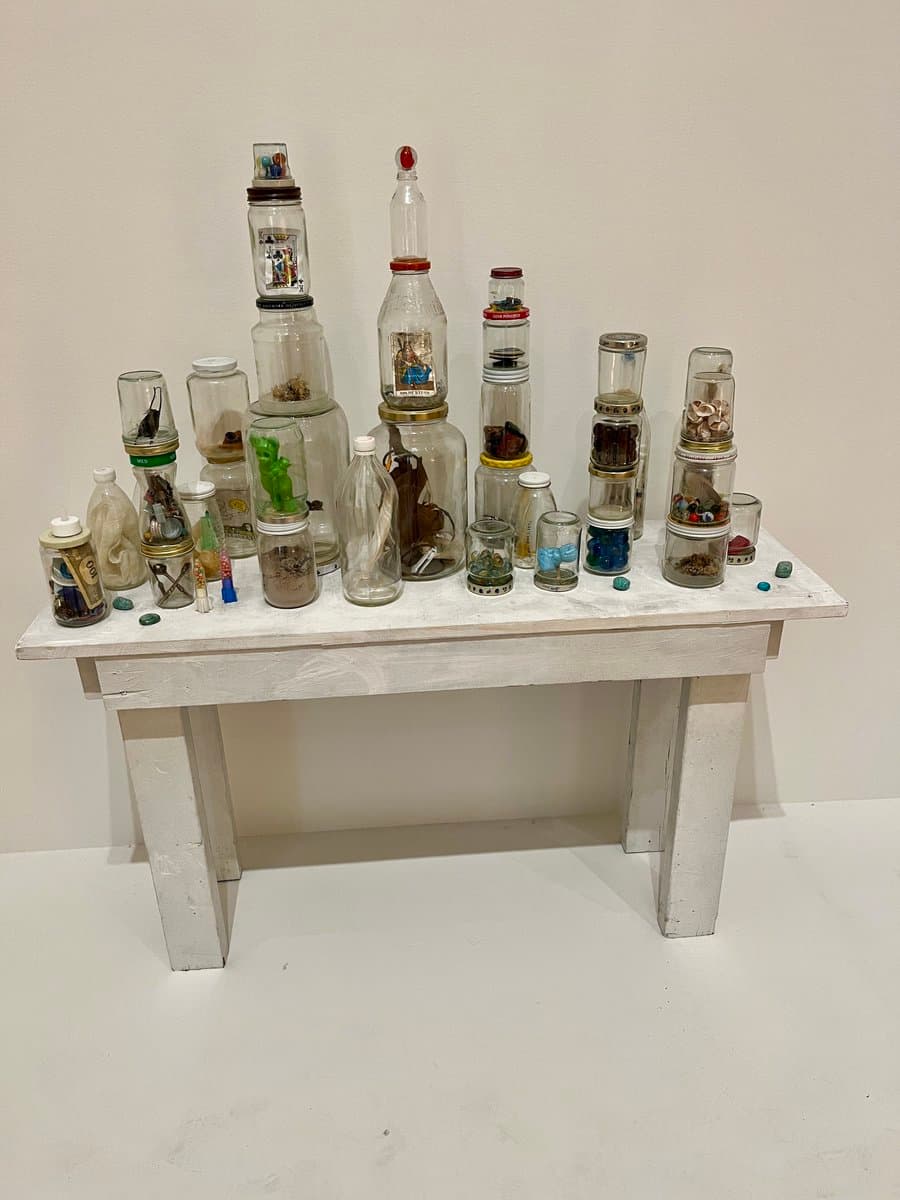A collection of glass bottles and jars of various sizes, filled with small objects and stacked in columns, displayed on a white table against a white wall.
