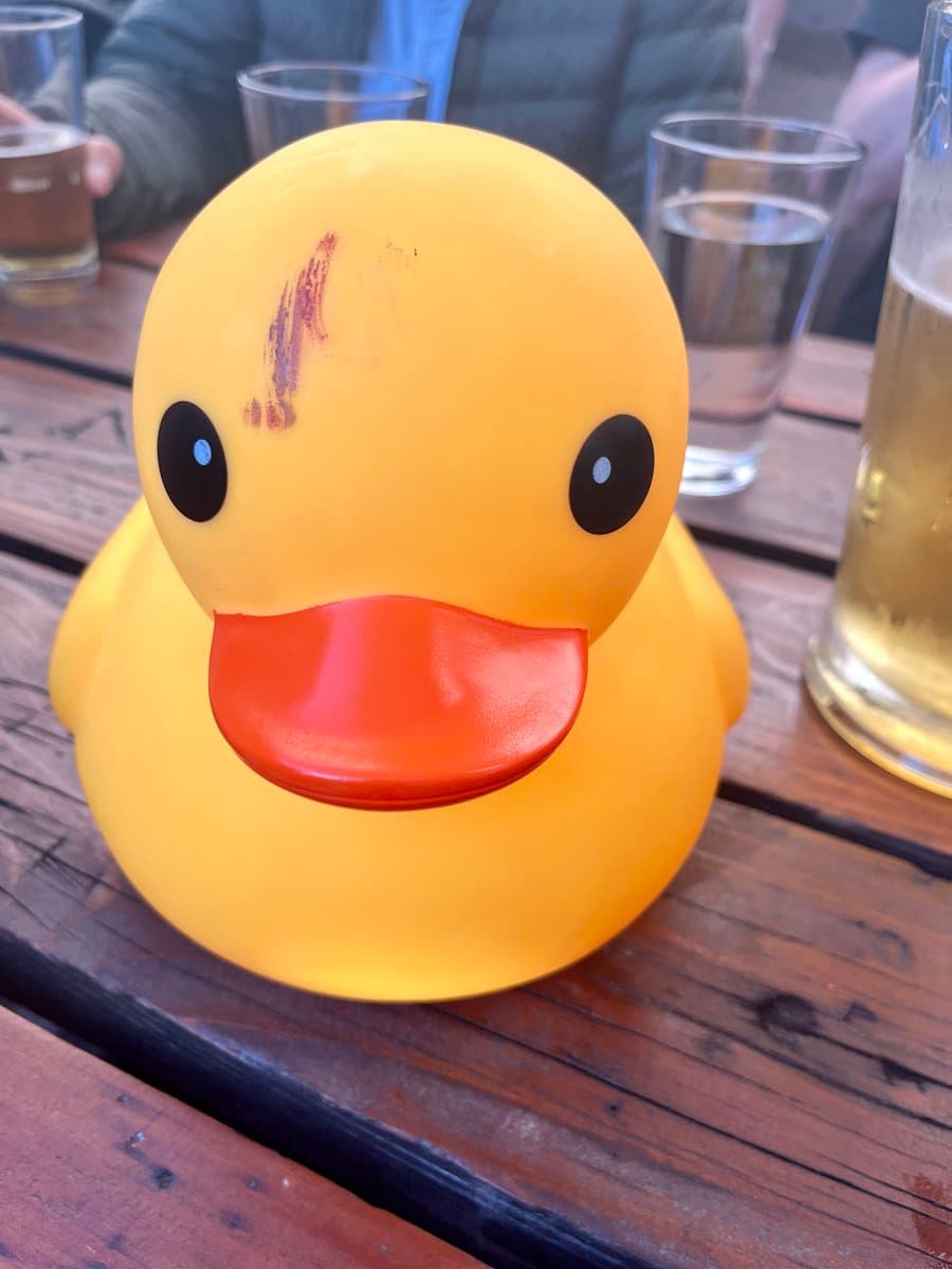 A close-up photograph of a yellow rubber duck with an orange beak sitting on a wooden table. The duck has a noticeable reddish-brown scratch or mark on the left side of its head. In the background, there are glasses containing beverages and a person wearing a dark jacket.