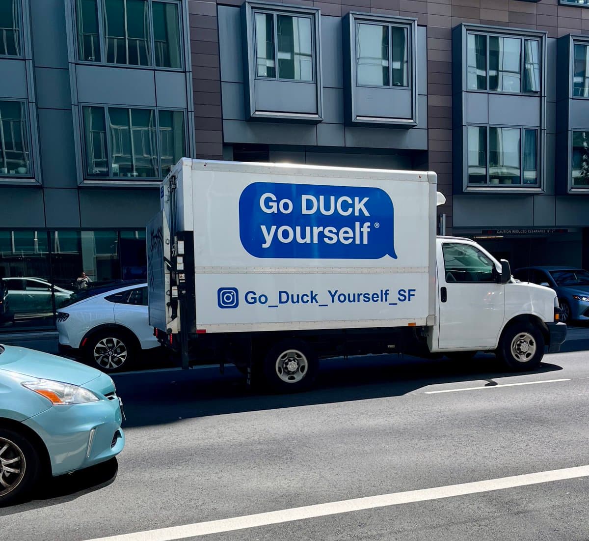 A white delivery truck is parked on a city street in front of a modern building, featuring a large blue speech bubble logo with the text 'Go DUCK yourself' and an Instagram handle 'Go_Duck_Yourself_SF' on its side. Other cars are visible in the foreground and background.