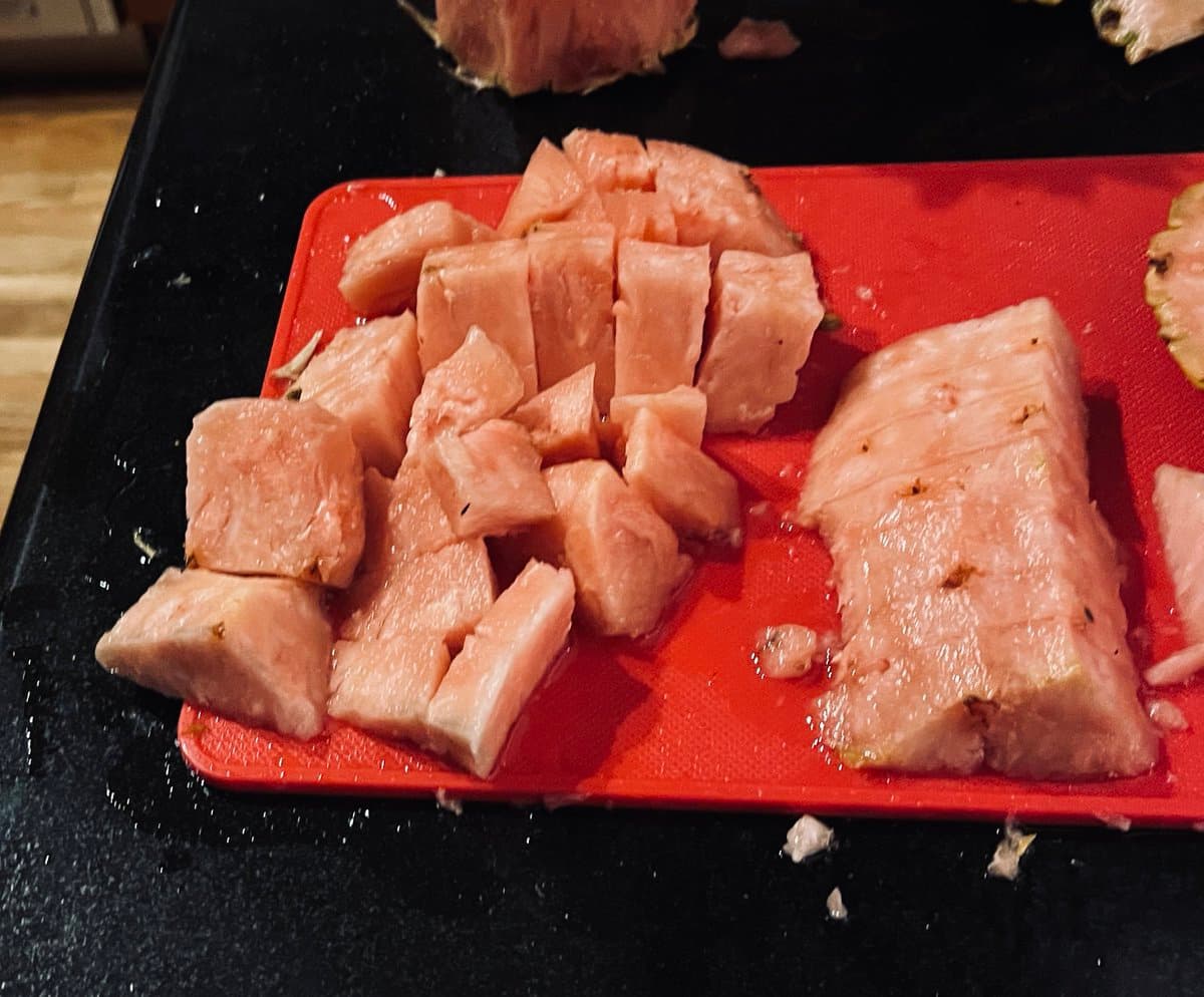 A close-up photograph of a red cutting board on a black countertop, holding chunks of a pink-fleshed fruit, likely a pink pineapple, that have been cut into cubes and slices.