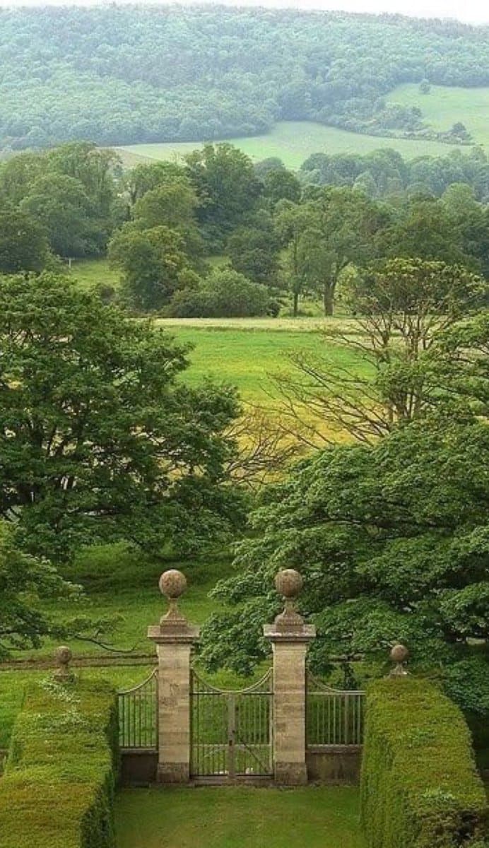 A scenic landscape photograph showing a stone gate with ornamental finials and metal fencing, set within a lush green garden with manicured hedges. Beyond the gate, the view opens up to rolling green fields, scattered trees, and a forested hill in the distance under an overcast sky.