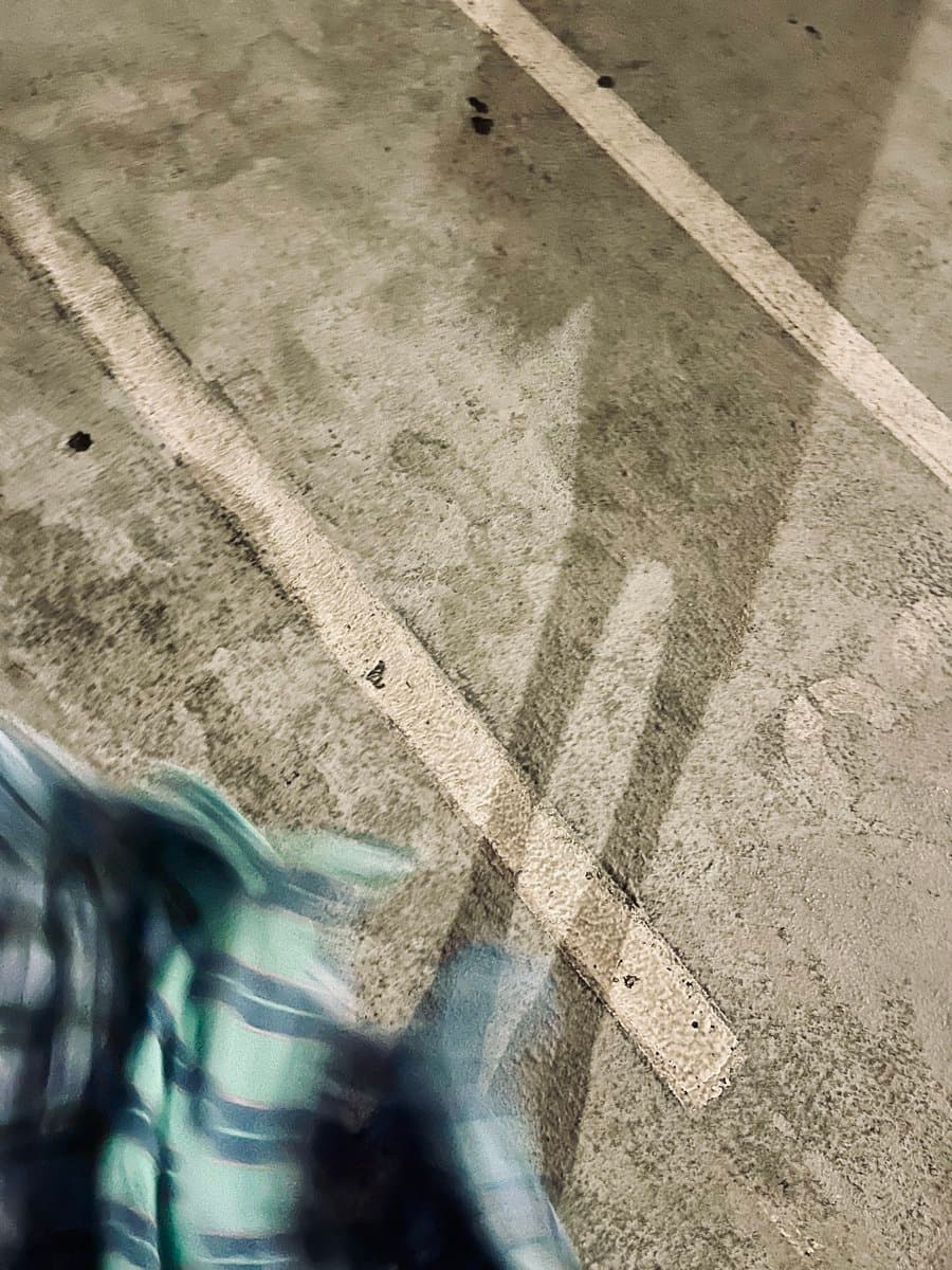 A close-up, slightly blurry photo of a concrete sidewalk with white painted lines, showing the shadow of a person's legs and feet. A portion of a plaid garment is visible in the lower left corner.