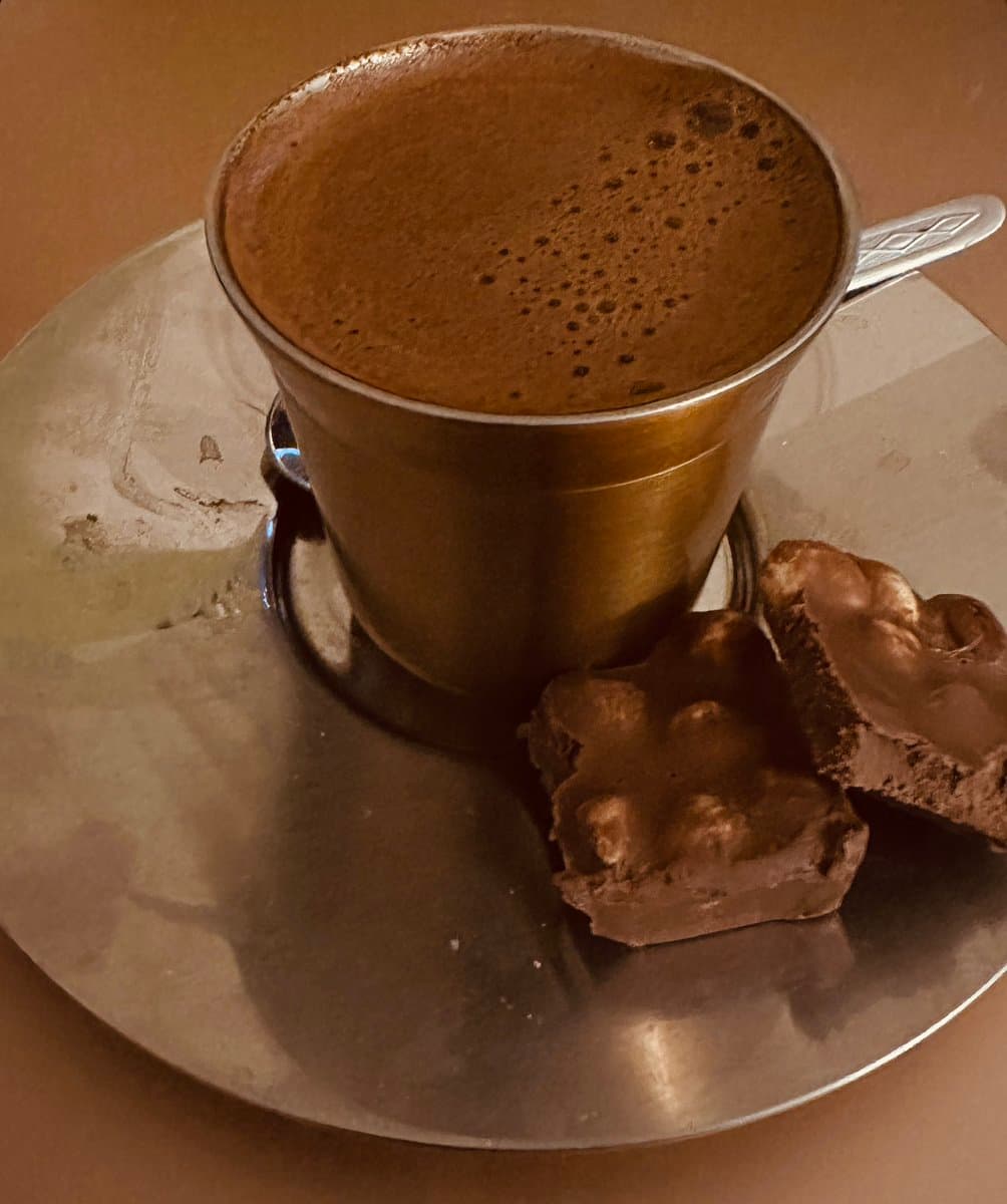 A close-up photograph of a tall, metallic cup filled with a frothy, brown hot beverage, likely a Turkish coffee or cappuccino, served on a silver platter. Two pieces of a chocolate-covered, textured confection, possibly a praline or nougat, sit on the plate next to the cup.