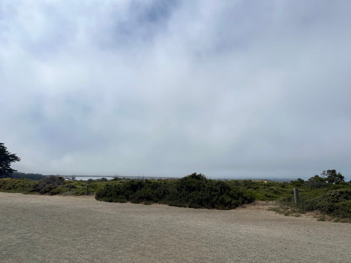 A landscape photograph showing a gravel path and low green bushes in the foreground, with a bridge barely visible in the distance through thick fog and clouds.