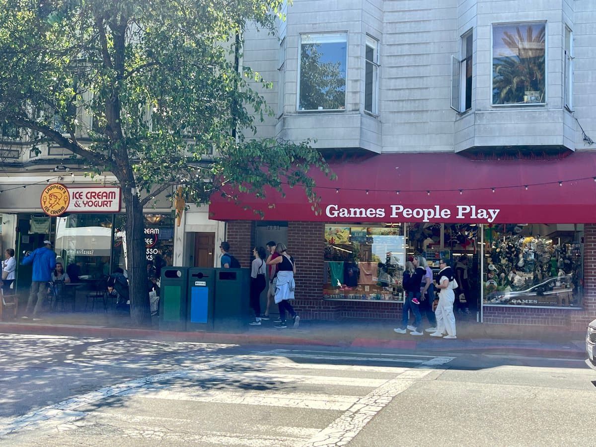 A street-level photograph of a storefront with a red awning reading 'Games People Play,' located next to an ice cream and yogurt shop. Several pedestrians are walking on the sidewalk in front of the stores, and a large tree partially obscures the view on the left.