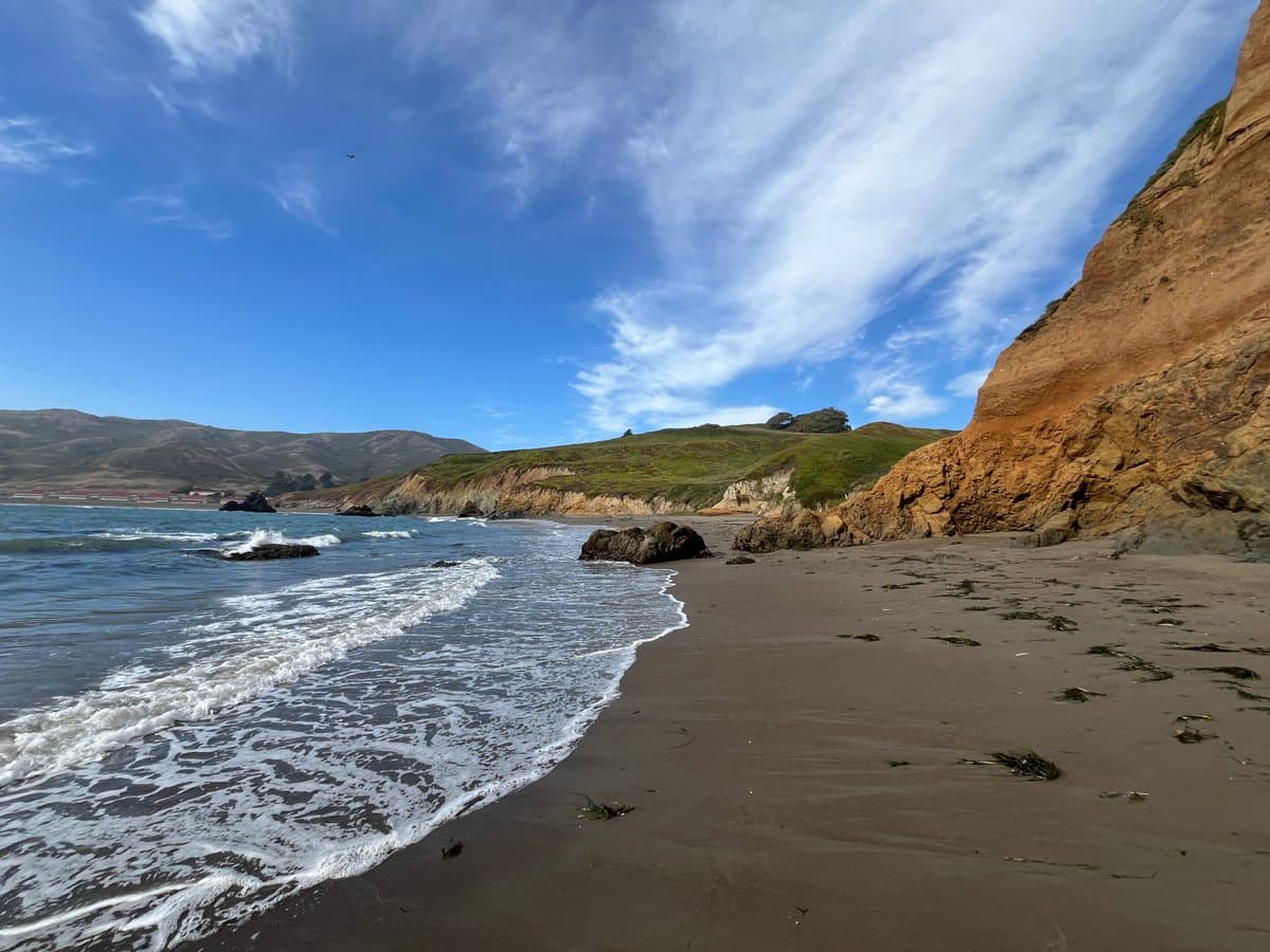 A wide landscape photograph of a quiet, sandy beach with gentle waves lapping at the shore. A steep, grassy cliff rises on the right side, while rolling hills are visible in the distance under a blue sky with wispy clouds.