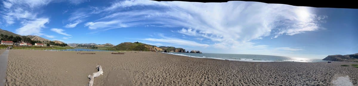 A wide panoramic photograph of a sandy beach with gentle waves, rolling hills, and a few buildings in the distance under a bright blue sky with wispy clouds.