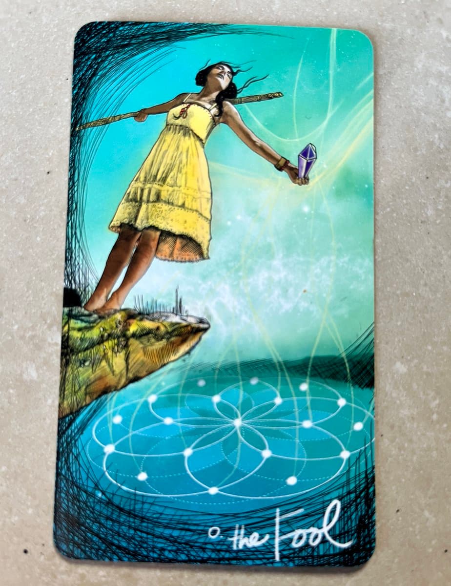 A photograph of a tarot card titled 'The Fool,' featuring an illustration of a woman in a yellow dress standing on a cliff's edge, holding a staff and a crystal, with a geometric flower-of-life pattern below. The card is placed on a light-colored, textured surface.