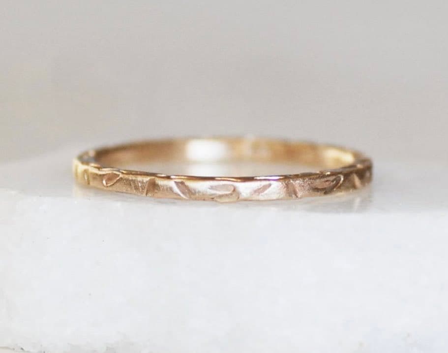A close-up photograph of a delicate, hammered gold band ring resting on a white surface.