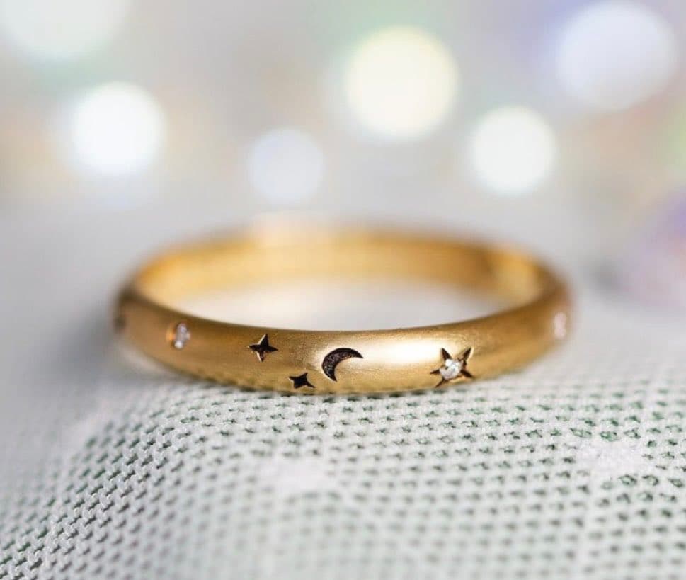 A close-up photograph of a delicate gold ring featuring engraved stars and a crescent moon, with small gemstones set into some of the stars. The ring rests on a textured white fabric against a softly blurred, bokeh-lit background.