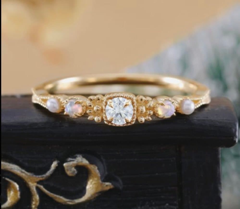 A close-up photograph of a delicate gold ring featuring a central round diamond and smaller opal-like stones, resting on a dark, ornate wooden stand.