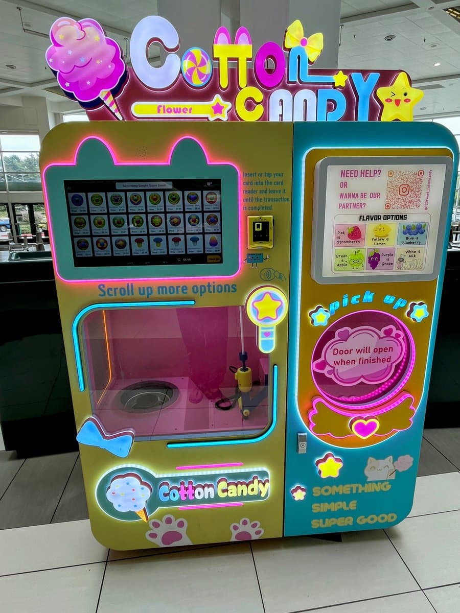 A brightly lit, pastel-colored vending machine for making cotton candy, featuring a claw-like mechanism and a digital selection screen. The machine is decorated with neon lights, stars, and cat-themed graphics.