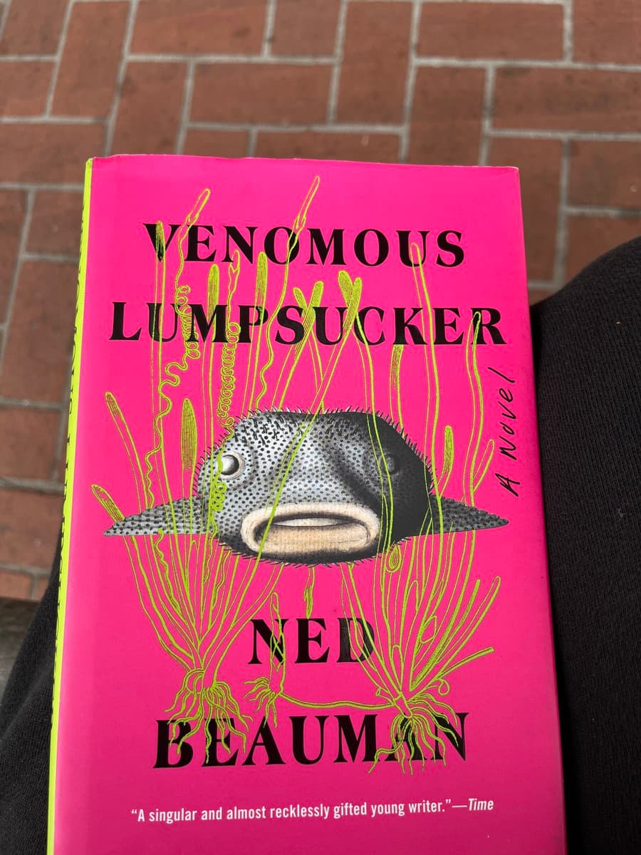 A photograph of the pink book cover for "Venomous Lumpsucker" by Ned Beauman, resting on a person's lap against a brick background. The cover features an illustration of a spiky, pufferfish-like creature with a human mouth, surrounded by green seaweed.