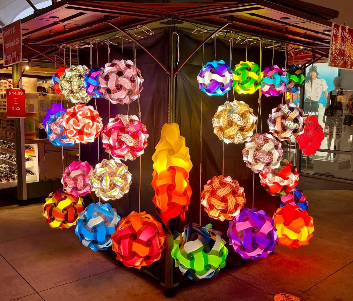 A display of colorful, illuminated paper lanterns hanging from a wooden canopy in a shopping area. The lanterns feature various vibrant patterns and shapes, including spherical and flame-like designs.