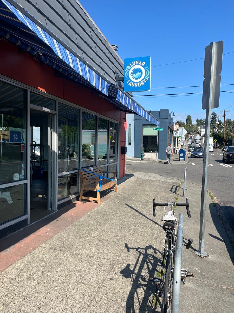 A street-level view of a storefront with a blue and white striped awning and a blue sign reading "LUNAR LAUNDRY". A wooden bench sits on the sidewalk in front of the shop, and a road bike is parked on a rack in the foreground.