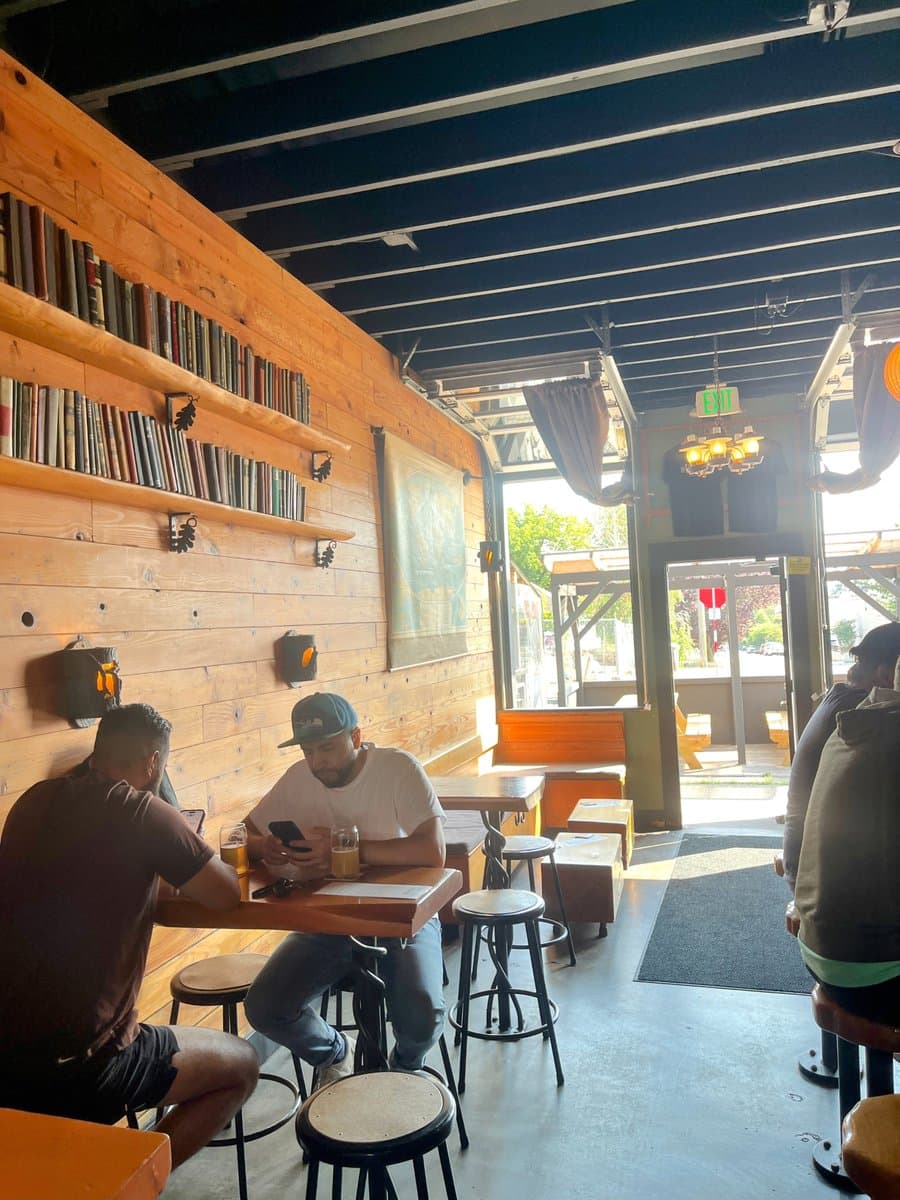 A photograph of the interior of a bar or restaurant featuring a rustic design with a wooden wall lined with bookshelves. Several people are seated at high-top tables and stools, with one man in a white t-shirt and cap looking at his phone. The space has an open ceiling with exposed beams and large windows looking out to a bright exterior.