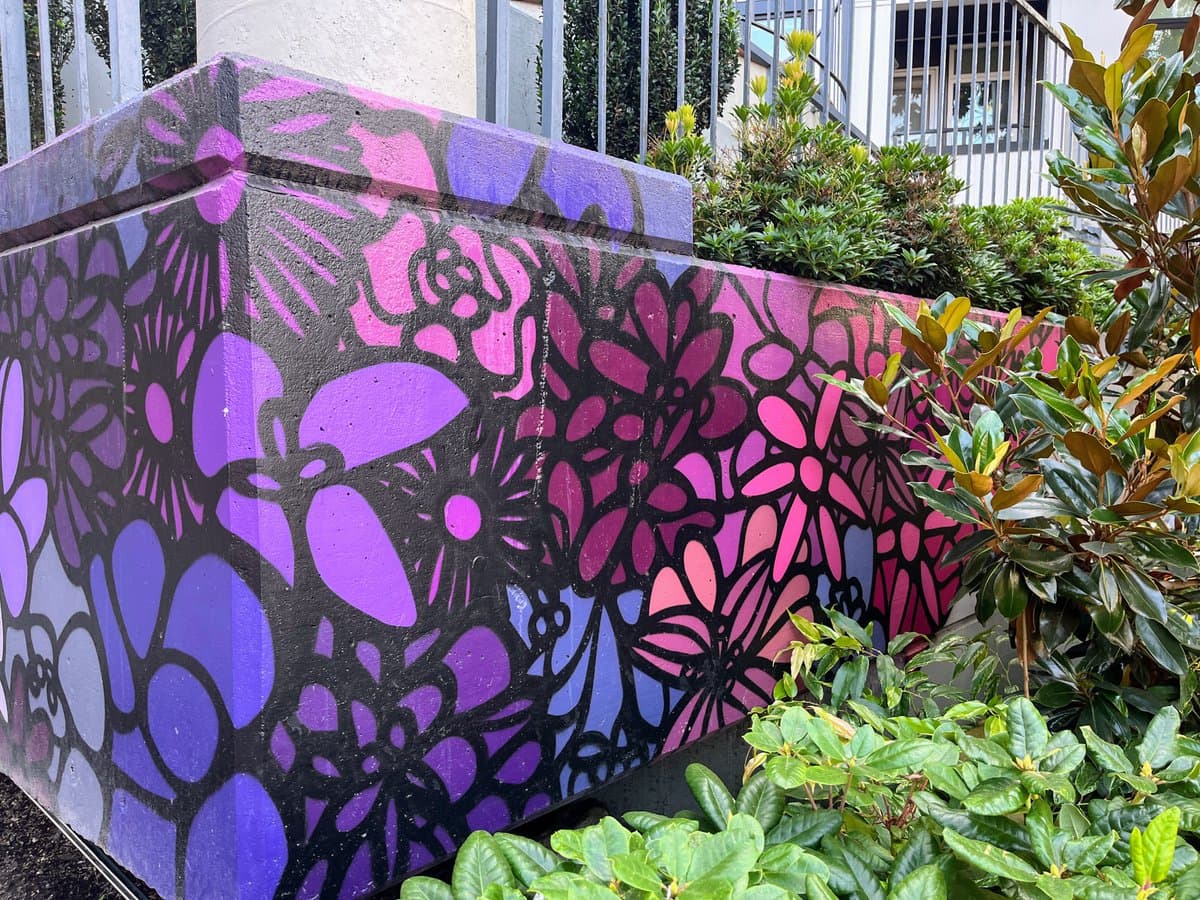 A close-up photograph of a concrete planter box decorated with a vibrant, stylized floral mural in shades of purple, pink, and black. The box is surrounded by green foliage, including a large leafy plant on the right, with a building and fence visible in the background.