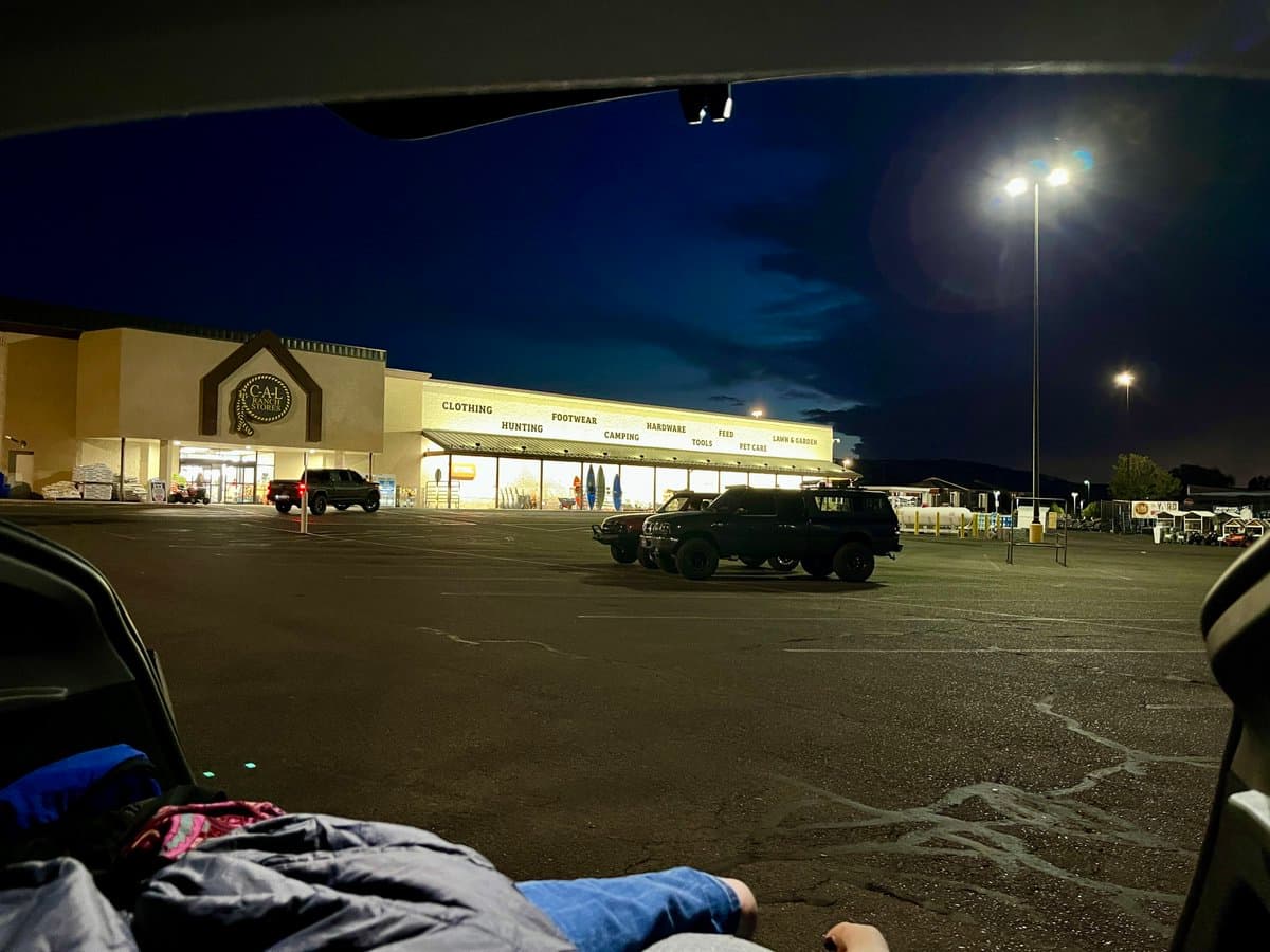 A first-person view from inside a vehicle looking out into a large, empty parking lot at night. In the background, a brightly lit Cabela's store is visible with several parked trucks in the foreground.