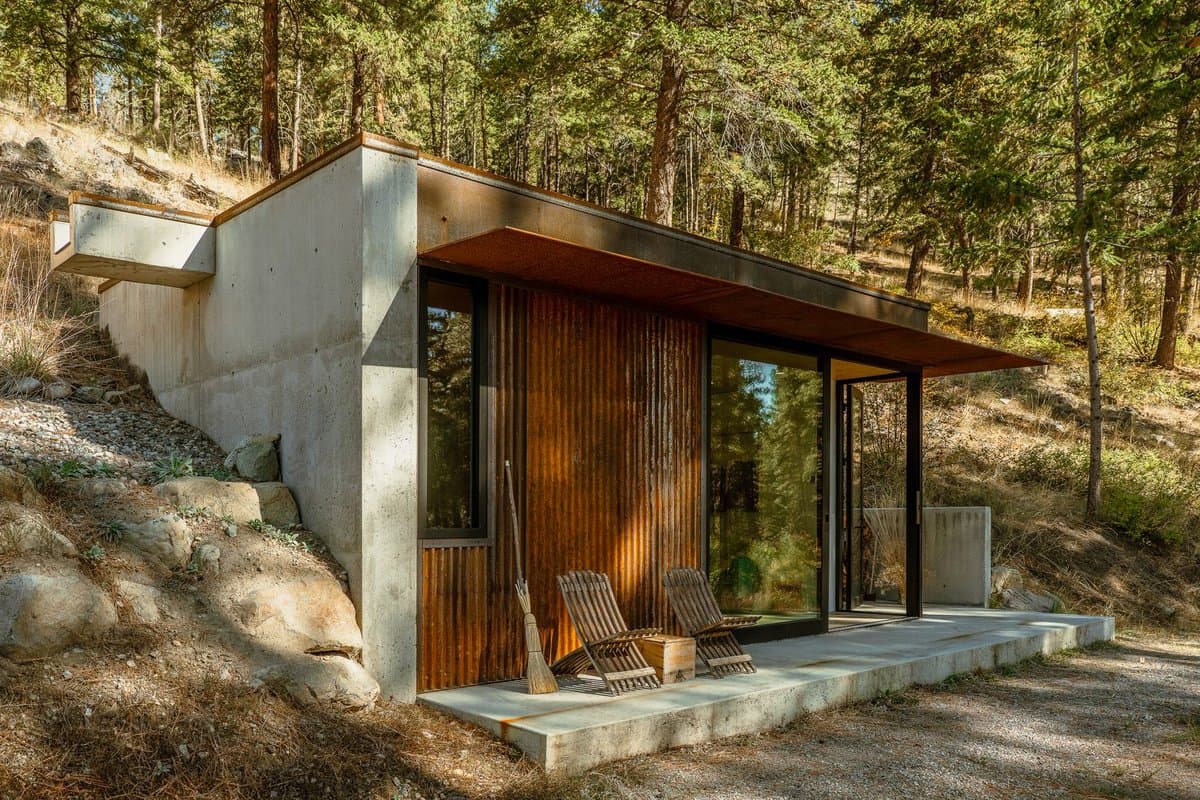 A modern cabin with concrete walls and a corrugated metal facade sits nestled into a forested hillside. The structure features large glass windows and a small concrete patio with two wooden chairs and a broom.
