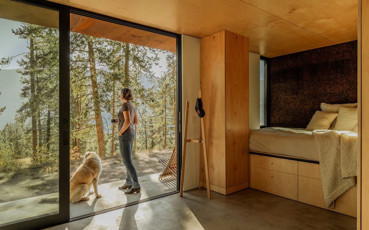 A woman and a dog stand on the concrete porch of a modern wooden cabin, looking out at a forested landscape through large glass sliding doors. The interior features warm wood paneling, a built-in bed with a cork headboard, and a minimalist design.