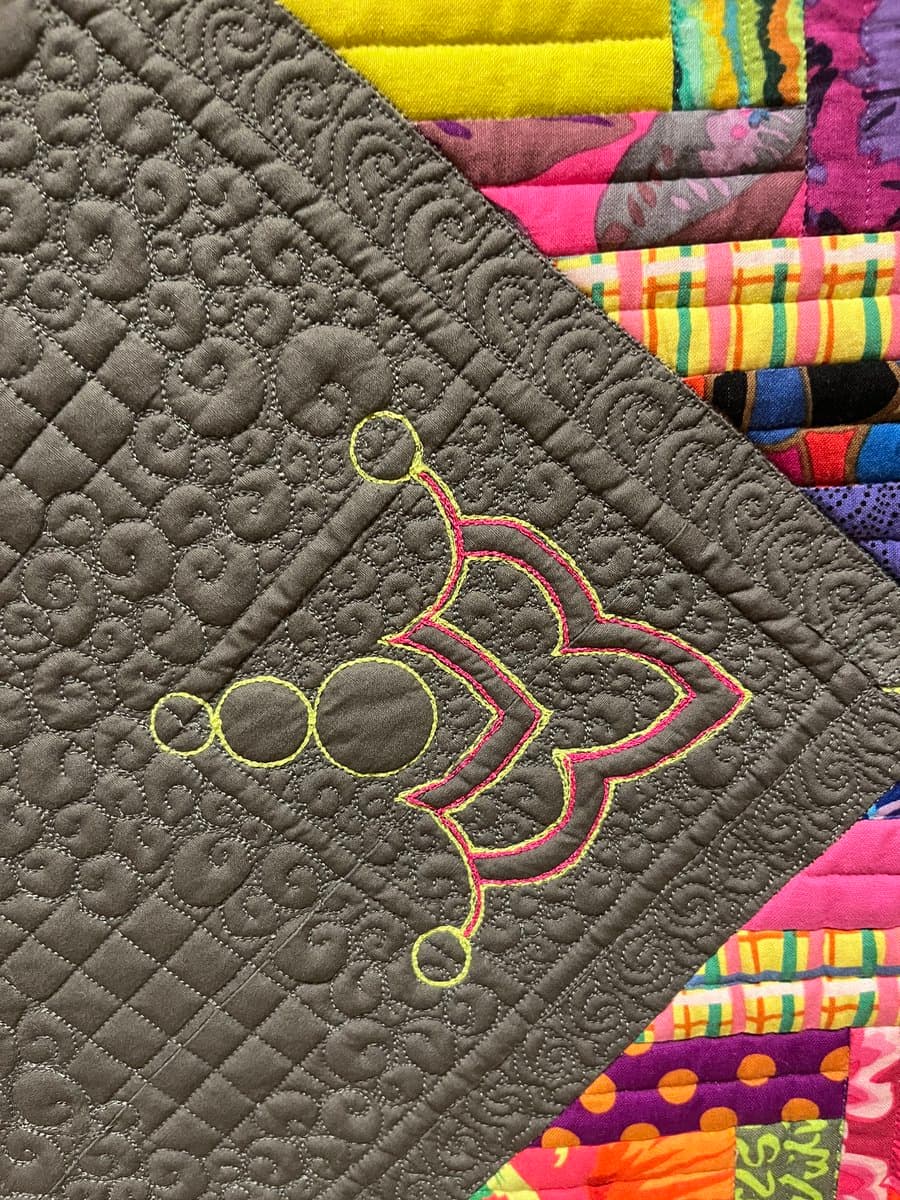 A close-up view of a quilt featuring a brown, densely stitched border with a decorative, embroidered motif in yellow and pink thread. The border frames a colorful patchwork of various patterned fabrics, including stripes, dots, and floral prints.
