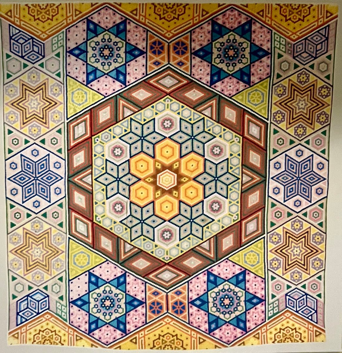 A colorful, geometric quilt featuring a large central hexagonal medallion surrounded by intricate star and cube patterns in a symmetrical layout. The design utilizes a vibrant palette of blues, yellows, pinks, and greens, showcasing detailed piecing and appliqué work. The quilt is displayed flat, likely on a wall or table, highlighting its complex craftsmanship.