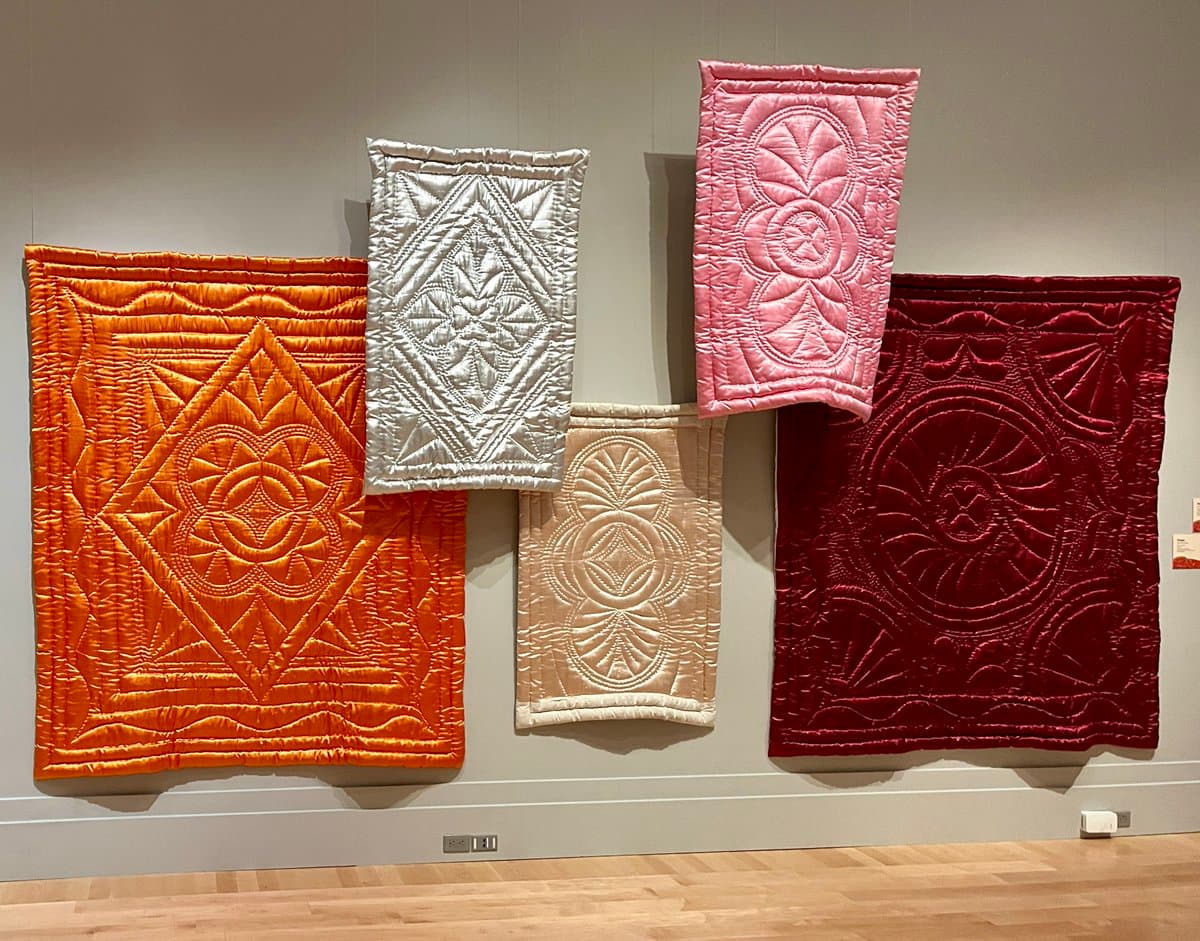 A photograph of five colorful, intricately quilted fabric panels hanging on a museum wall. The pieces feature various geometric and floral patterns in shades of orange, white, pink, beige, and red.