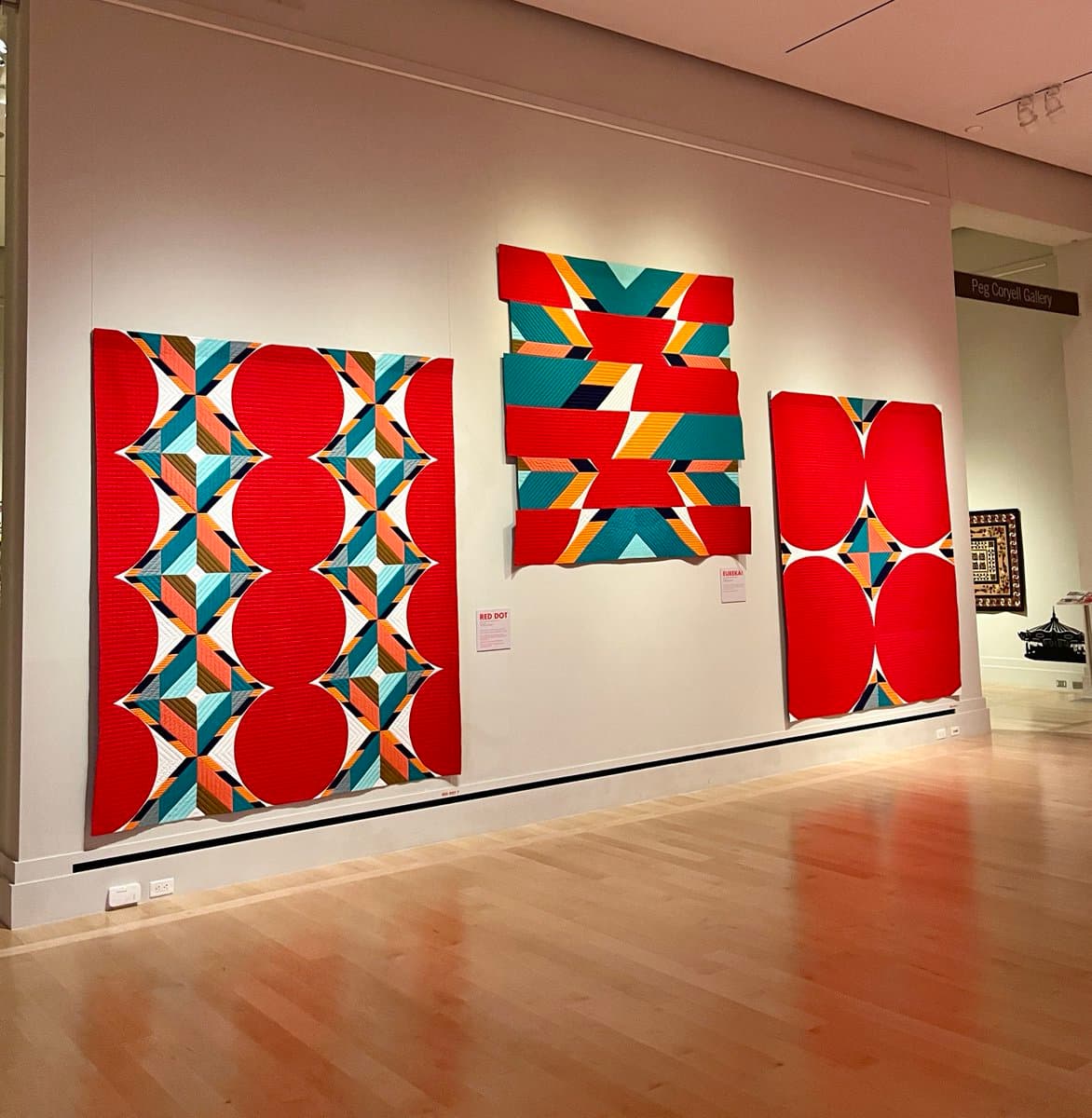 A photograph of a museum gallery featuring three large, colorful geometric quilts hanging on a white wall. The quilts feature bold red, teal, orange, and white patterns. A sign in the background reads "Peg Coryell Gallery".