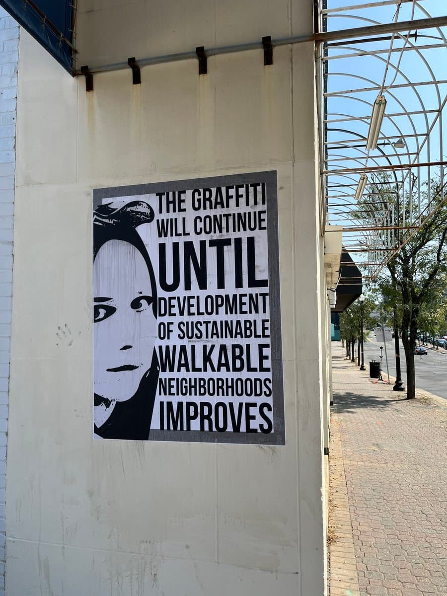 A black-and-white street art poster is affixed to a white construction hoarding on a city sidewalk. The poster features a stylized portrait of a person and bold text advocating for sustainable urban development.