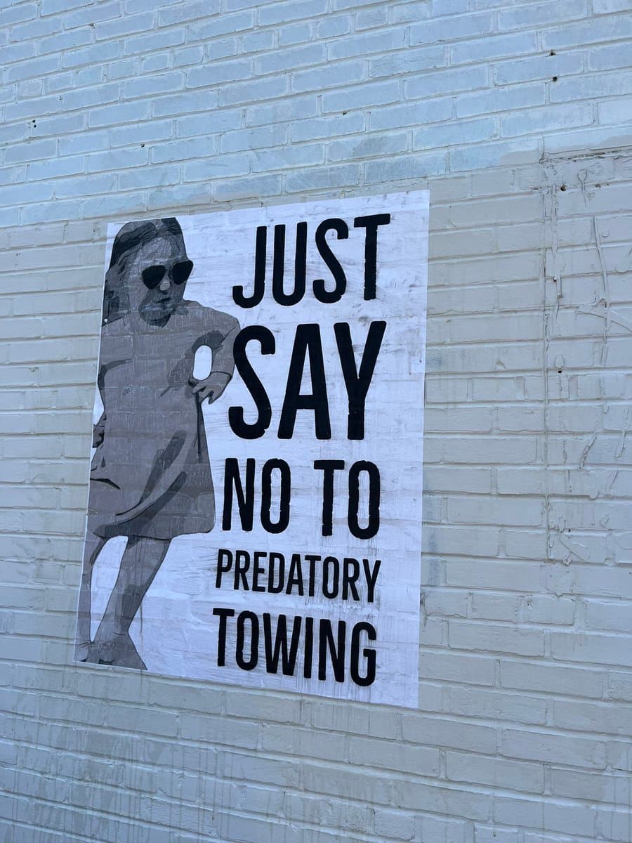A black-and-white protest poster is taped to a white brick wall, featuring a stylized illustration of a person wearing sunglasses and a coat. The poster displays large, bold text reading 'JUST SAY NO TO PREDATORY TOWING.'