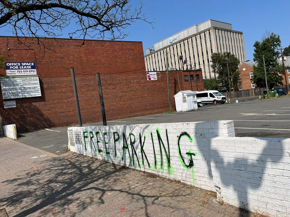 A street-level photograph of a parking lot on a sunny day, featuring a low white brick wall in the foreground with the words 'FREE PARKING' spray-painted in black and green. In the background, there is a red brick building with an 'OFFICE SPACE FOR LEASE' sign, a white van, and a tall office building.