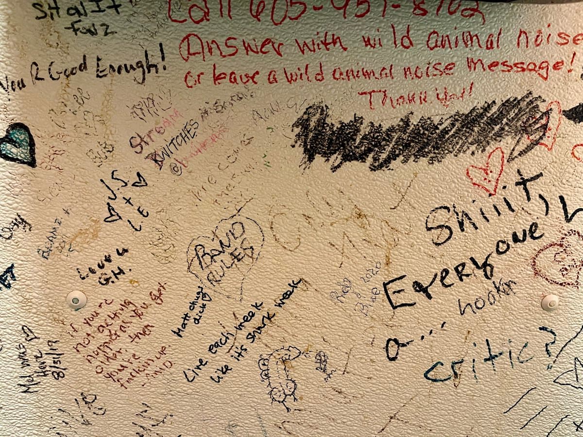 A close-up photograph of a textured, off-white surface covered in handwritten notes, doodles, and signatures in red and black ink. The image captures a chaotic collage of messages, including a phone number, instructions, and various signatures, resembling a guest book or a wall of feedback.