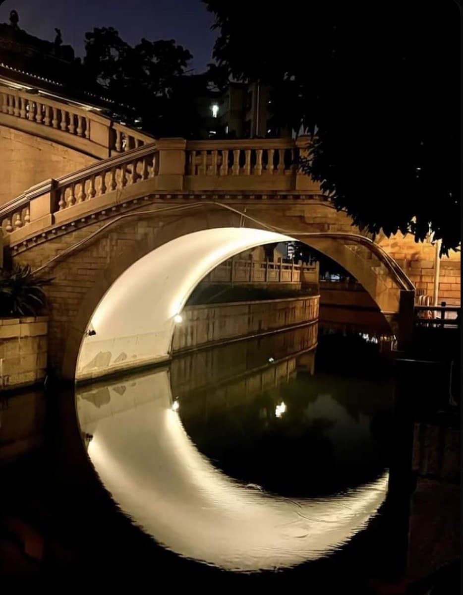 A nighttime photograph of a stone bridge over a canal, where the illuminated arch and its reflection in the water form the shape of a crescent moon. Silhouetted trees and buildings are visible in the background against a dark sky.