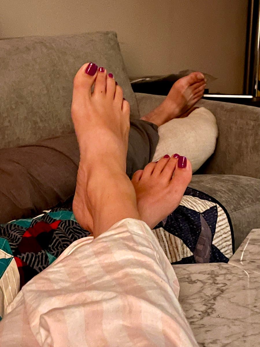A first-person view of a person's bare feet with dark red nail polish resting on a couch, with another person's bare feet visible in the background. The person in the foreground is wearing light-colored striped pants and is sitting on a patterned blanket.