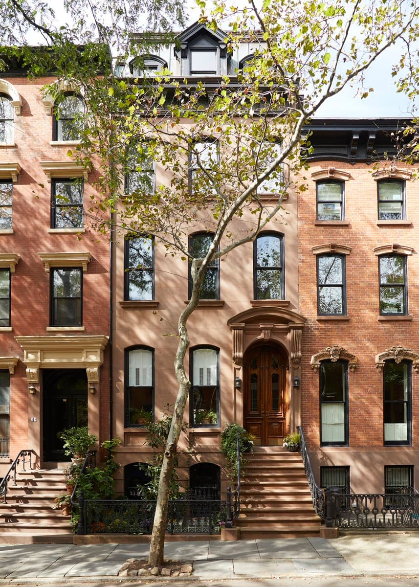 A street-level photograph of a row of classic, multi-story brick and stucco townhouses with ornate architectural details, front stoops, and black iron railings. A slender tree stands in the foreground on the sidewalk, partially obscuring the central building.