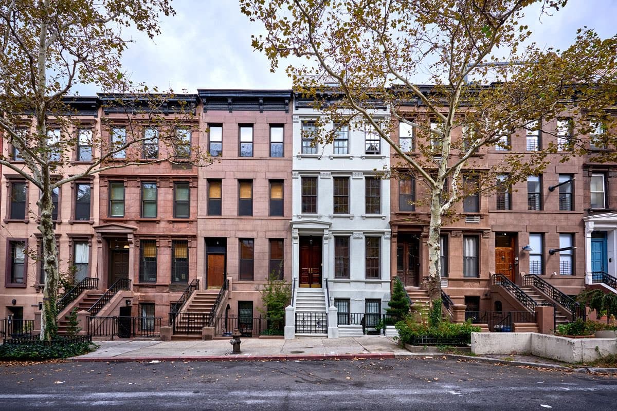 A street-level photograph showing a row of multi-story brick townhouses in various shades of brown, pink, and white, featuring stoops and black railings. Leafless trees line the sidewalk in front of the buildings, and a wet asphalt street is visible in the foreground.