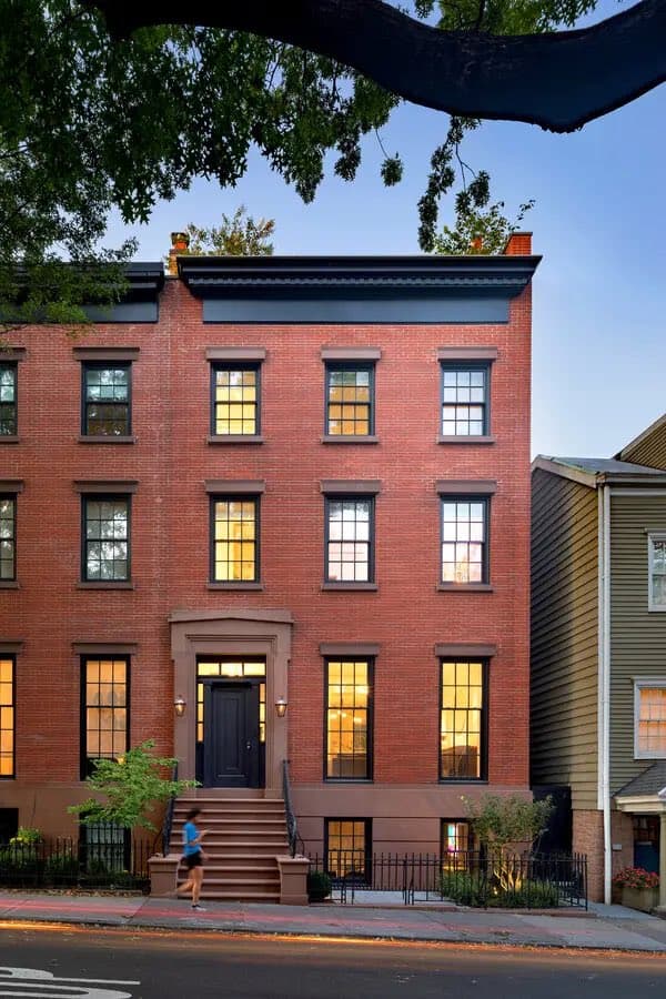 A photograph of a three-story red brick townhouse with a flat roof, featuring multiple windows with warm interior lighting and a central front entrance with steps. A person is walking up the steps, and the building is framed by tree branches overhead and a neighboring house to the right.