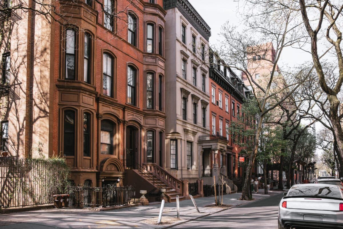 A street-level photograph of a row of historic brownstone townhouses in a city, featuring red brick and stone facades, with a white car parked on the street and bare trees lining the sidewalk.