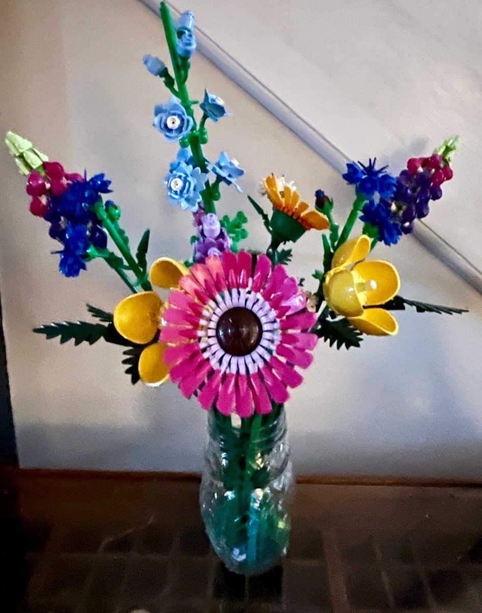 A photograph of a colorful bouquet of flowers constructed from plastic building blocks, arranged in a clear glass vase on a wooden surface.
