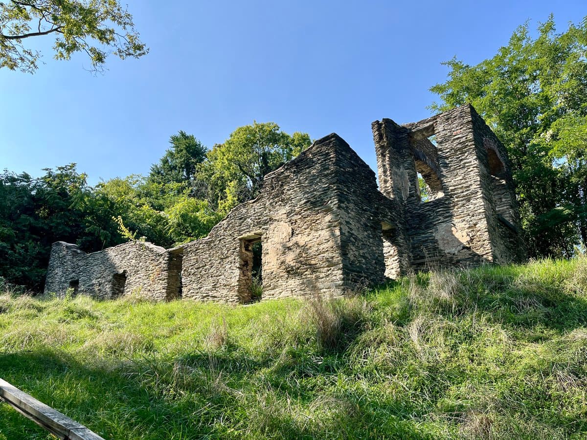 A photograph showing the stone ruins of a historic building on a grassy hillside, surrounded by trees under a clear blue sky.