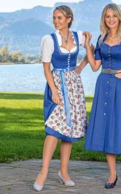 Two women wearing traditional blue dirndls stand outdoors on a paved path near a lake with mountains in the background. The woman on the left wears a dirndl with a floral apron, while the woman on the right wears a solid blue dirndl with a silver belt.