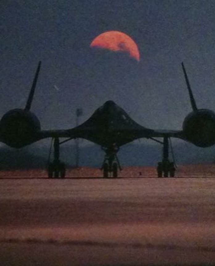 A low-angle photograph of a large, dark military aircraft parked on a tarmac at dusk, with a large, reddish moon rising in the sky behind it. The image has a grainy, vintage quality.