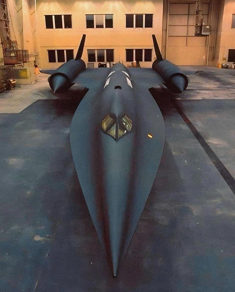 A front-facing, low-angle photograph of a dark, stealthy military aircraft with a pointed nose and twin vertical stabilizers, parked on a concrete tarmac in front of a large hangar building.