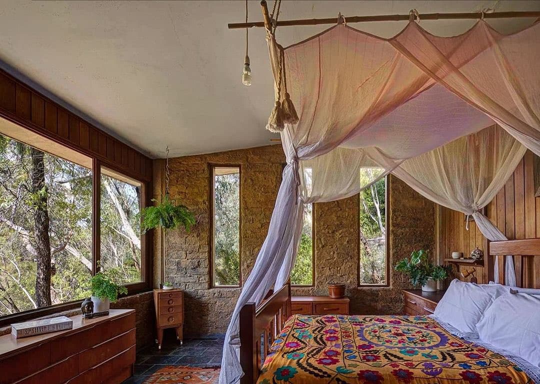 A rustic bedroom featuring a wooden bed with a colorful, patterned quilt and a sheer white canopy draped from a bamboo pole. The room has stone walls, large windows looking out onto a forest, and wooden furniture, creating a cozy, bohemian atmosphere.