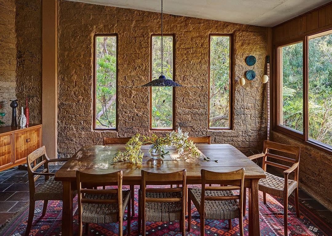 A rustic dining room with textured, earthen walls and a large wooden table surrounded by woven chairs. The space features tall, narrow windows looking out onto greenery, a patterned rug on the floor, and a hanging pendant light over the table.