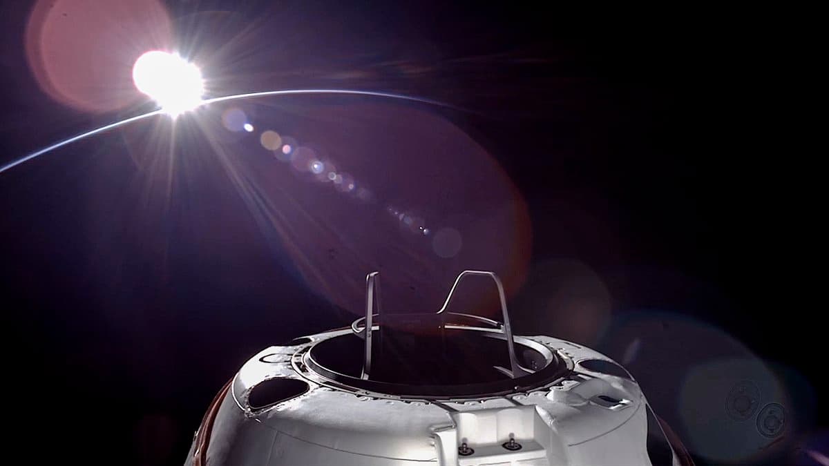 A view from space showing the top of a white spacecraft capsule with a docking mechanism, set against the dark background of space with the sun shining brightly over the Earth's horizon.