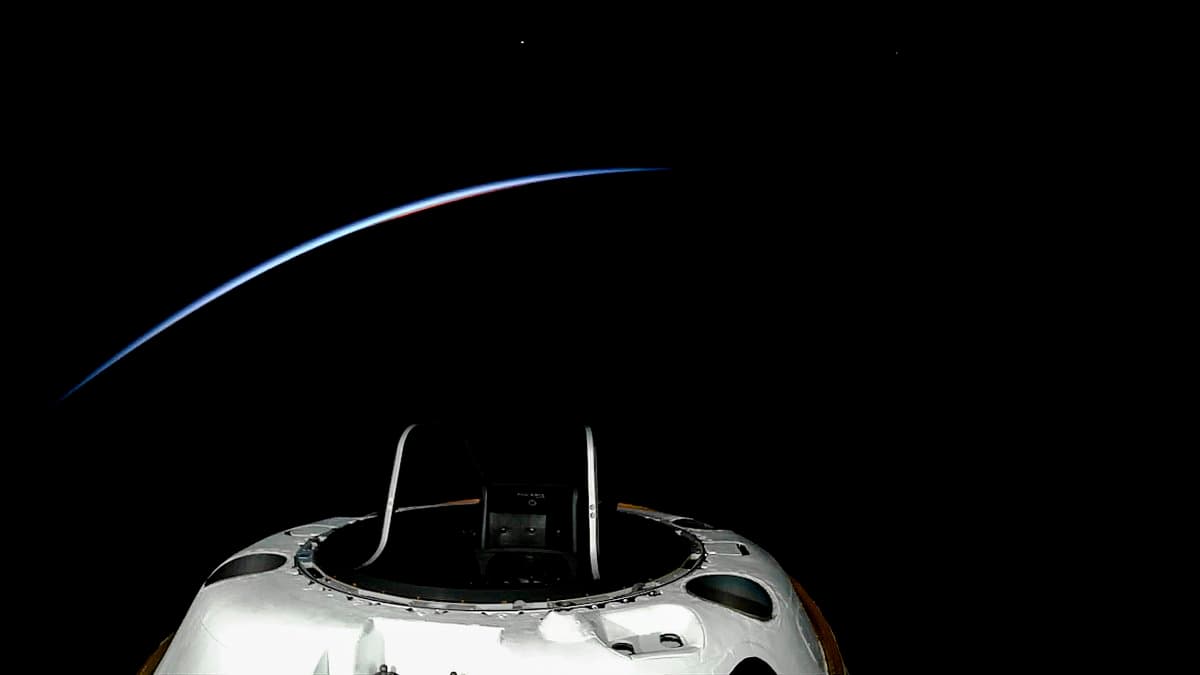 A photograph taken from space showing the top of a white spacecraft module against the blackness of space, with the thin, glowing blue and orange curve of Earth's atmosphere visible in the background.