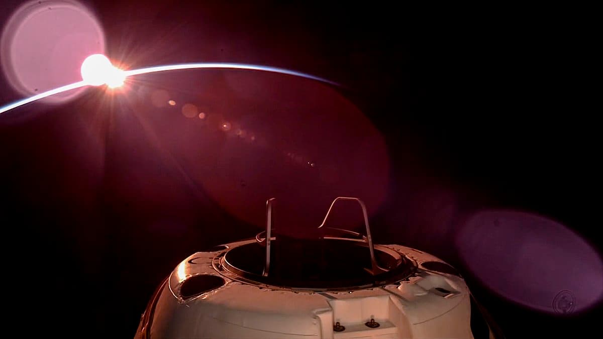 A view from space showing the top of a white spacecraft module with a docking mechanism, set against the dark background of space with the sun visible on the horizon of a planet.